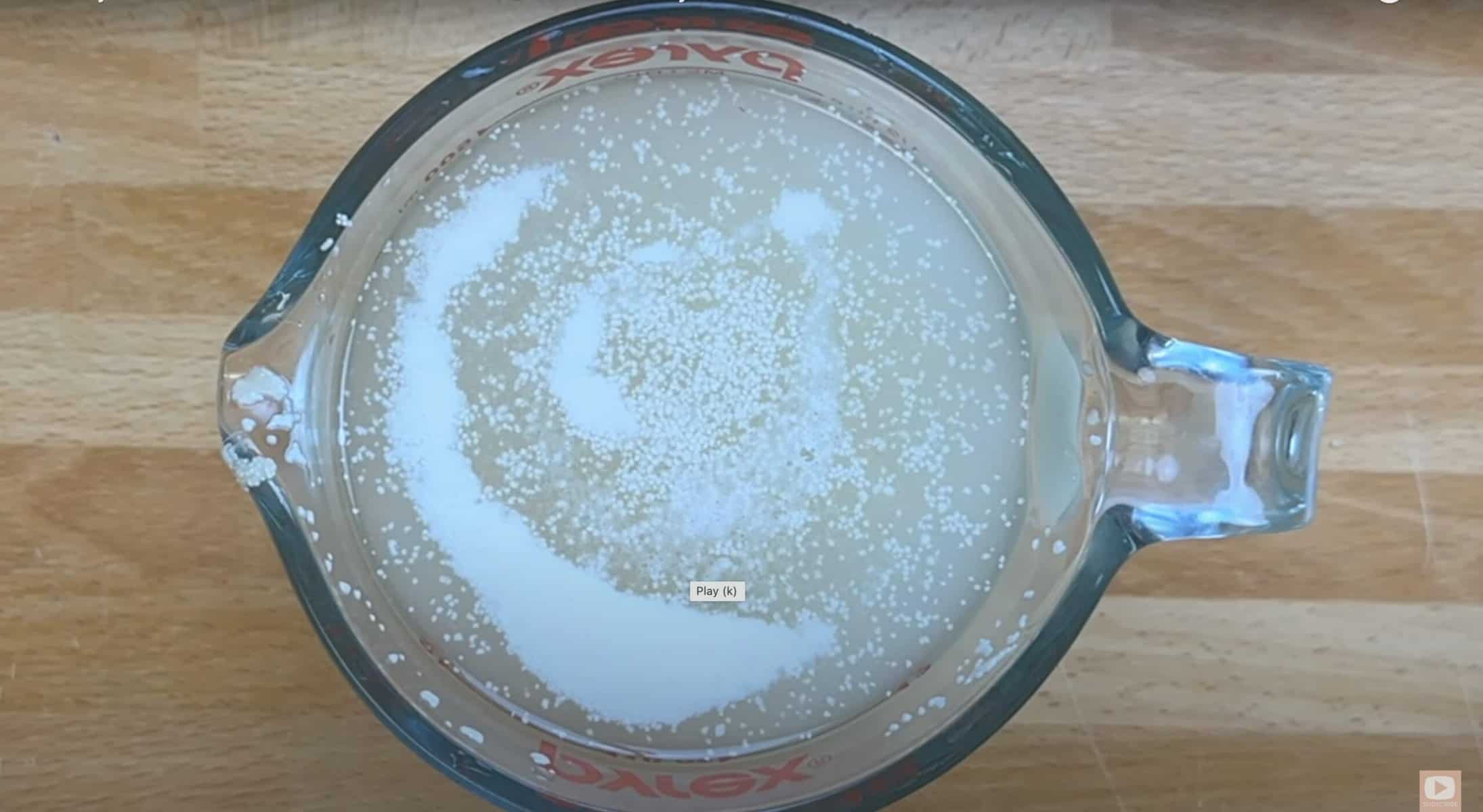 Yeast blooming in warm water inside a Pyrex measuring cup on a wooden surface.